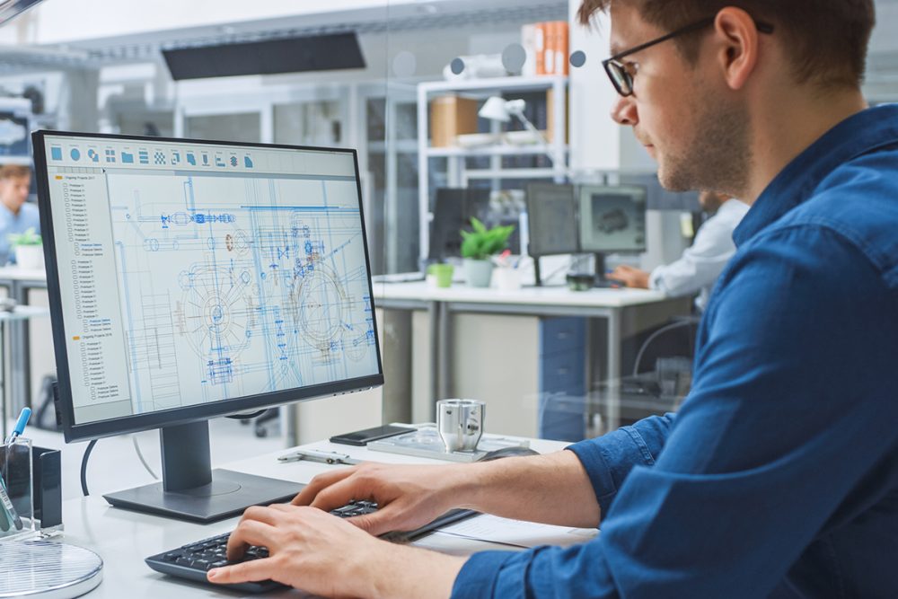 Over the Shoulder Shot of Engineer Working with CAD Software on Desktop Computer, Screen Shows Technical Drafts and Drawings. In the Background Engineering Facility Specialising on Industrial Design